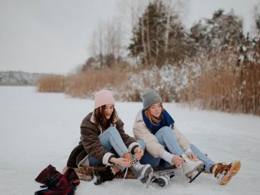 Two women sitting on a frozen lake tying ice skates, enjoying winter leisure outdoors.
