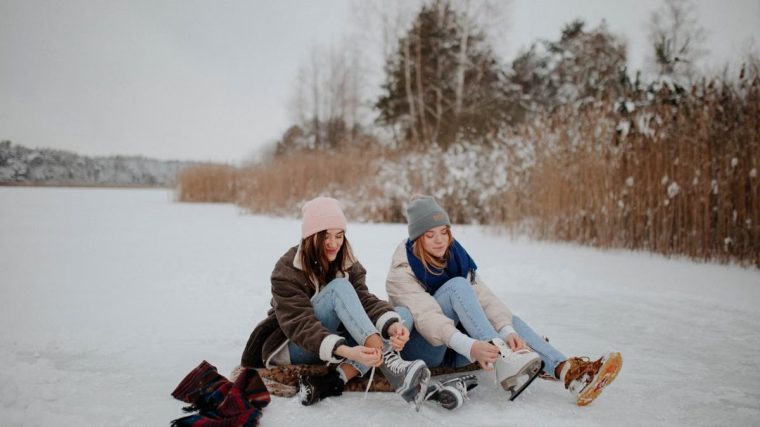 Two women sitting on a frozen lake tying ice skates, enjoying winter leisure outdoors.